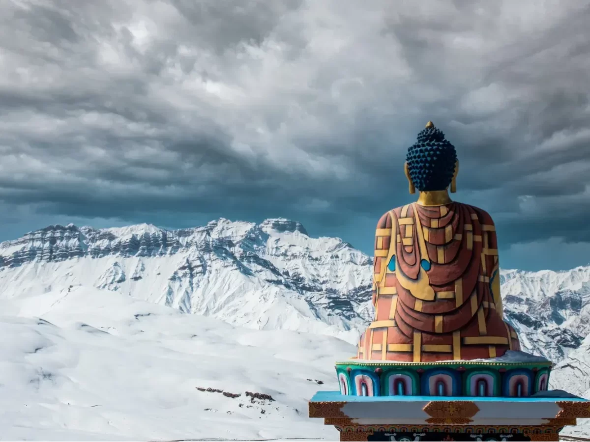 Langza Village near Kaza Spiti winter serenity, featuring giant Buddha statue overlooking snow-clad mountains cloudy skies dramatic Himalayan backdrop, perfect Himachal Pradesh tour packages.