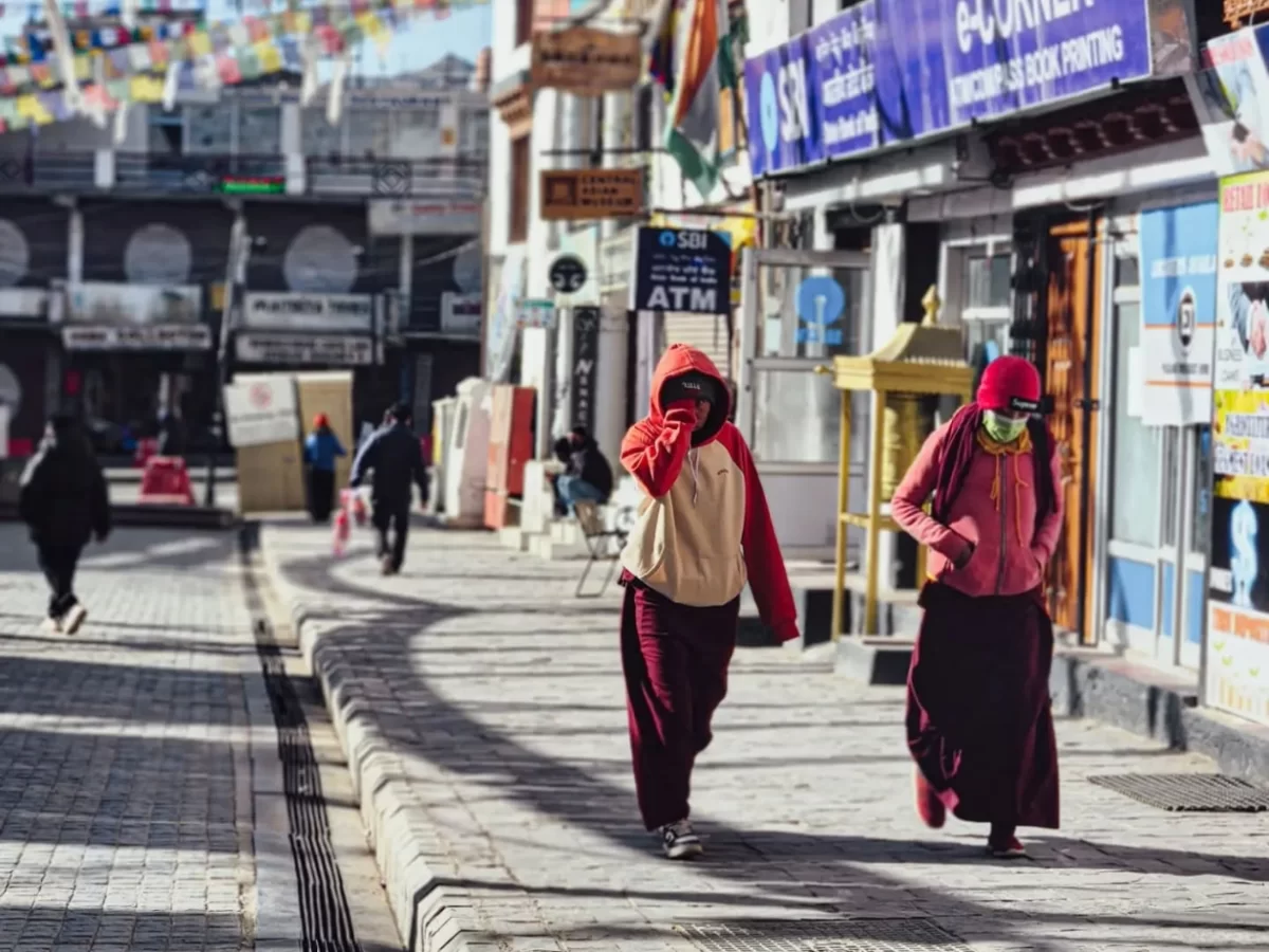 Two masked monks in maroon robes walking Leh Market street under prayer flags near ATM and shops during sunny day, perfect India tour package