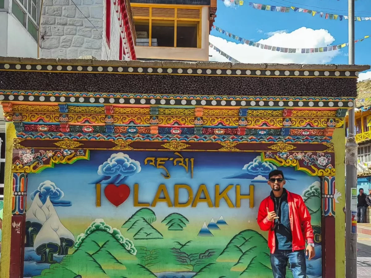 Vibrant I Love Ladakhh billboard with Tibetan script, mountains backdrop, man posing in red jacket amid prayer flags near Leh Market, perfect India tour package. 