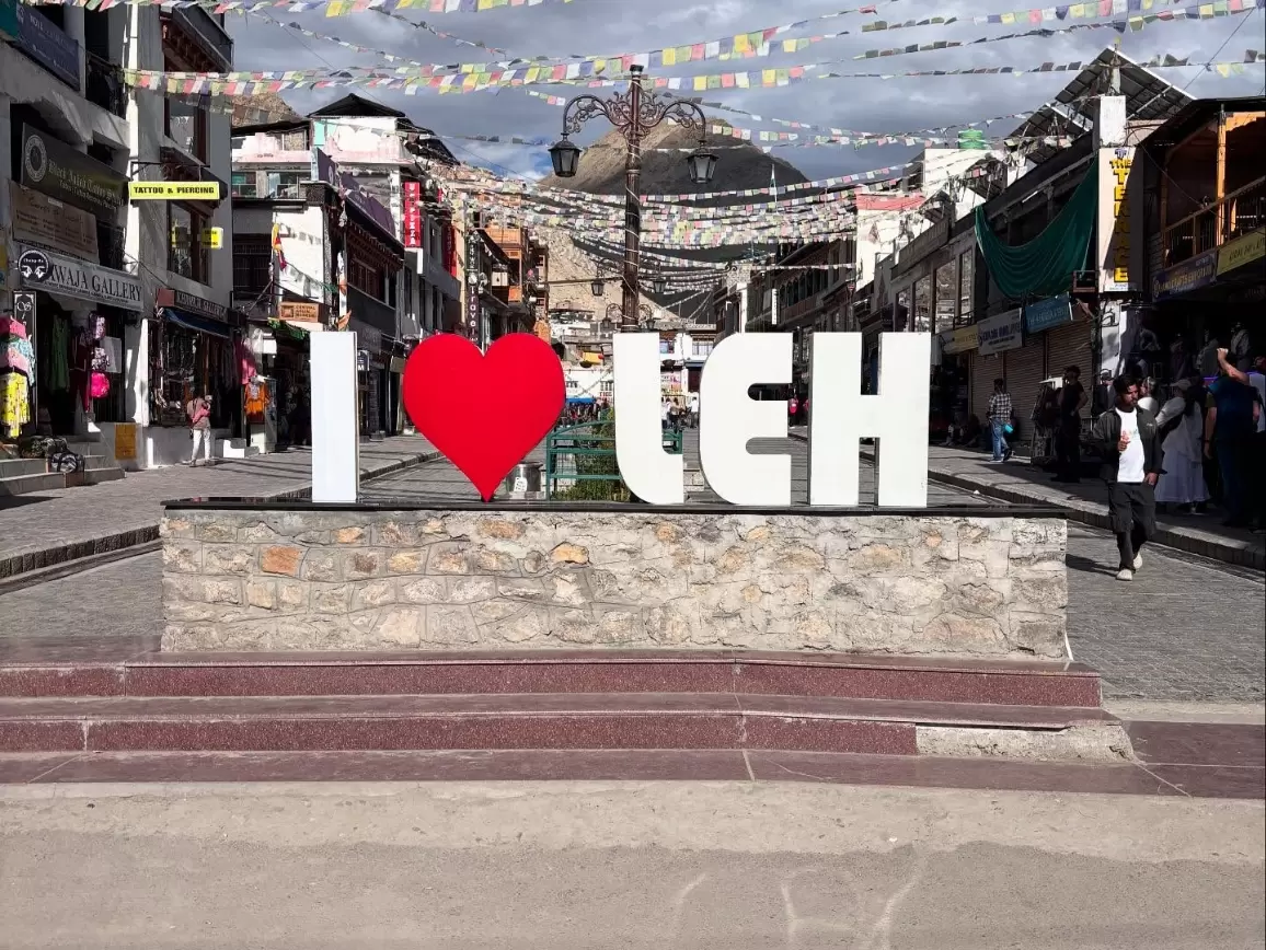 Iconic I Love Leh sign with large white letters and red heart in Leh Market under prayer flags, bustling shops backdrop, perfect India tour package.