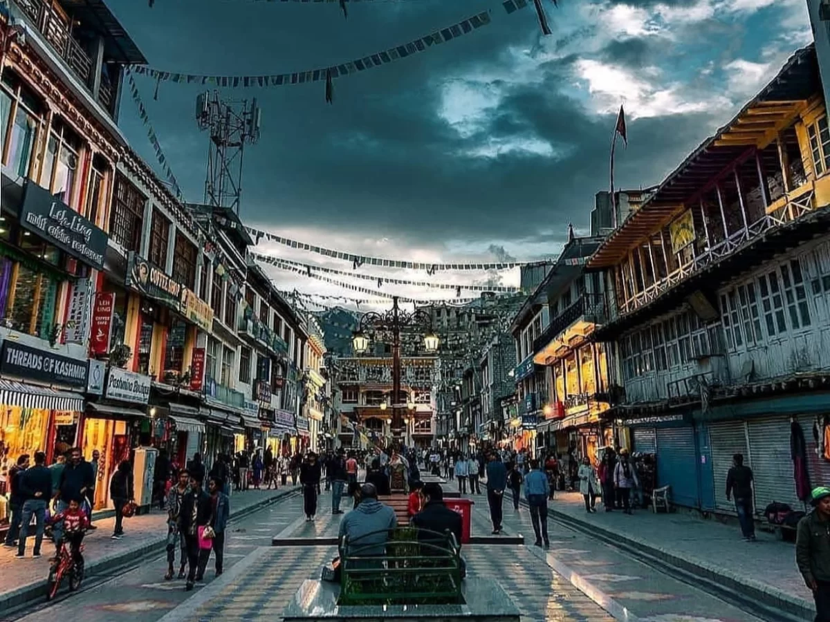 Bustling Leh Market street under dramatic cloudy sky with prayer flags, crowded shops and diverse pedestrians, perfect India tour package in Ladakh.