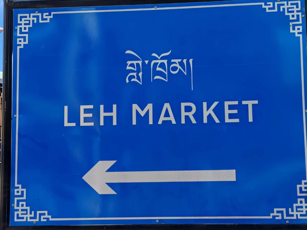 Blue signboard for Leh Market with Tibetan script, English text and left arrow, showcasing Ladakhi bilingual signage, perfect India tour package.