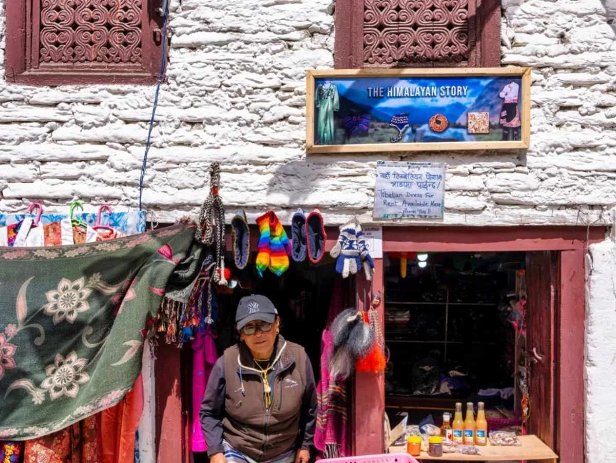 Ladakhi woman at The Himalaya Story shop in Leh Market displaying pashmina shawls, colorful scarves, handicrafts amid traditional white-washed building, perfect India tour package. 