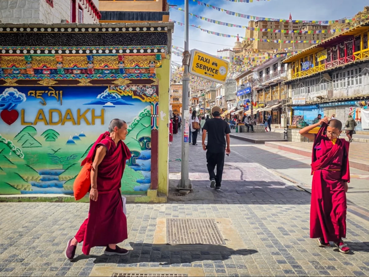 Monks in maroon robes passing taxi service stand amid prayer flags and colorful murals in Leh Market street, vibrant Himalayan culture, perfect India tour package. 