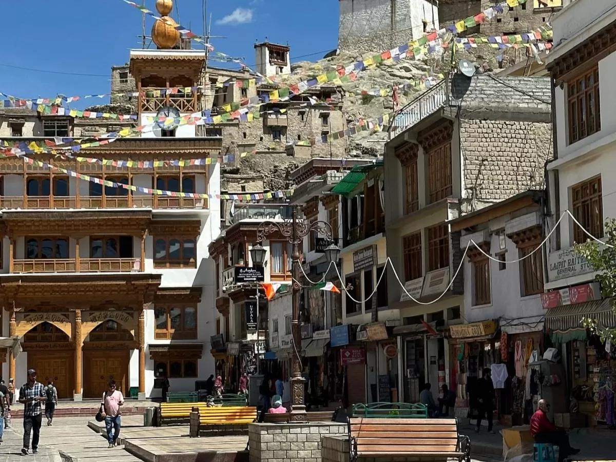 Vibrant Leh Market square with prayer flags, traditional Ladakhi architecture, golden stupa against blue sky and distant hills, perfect India tour package