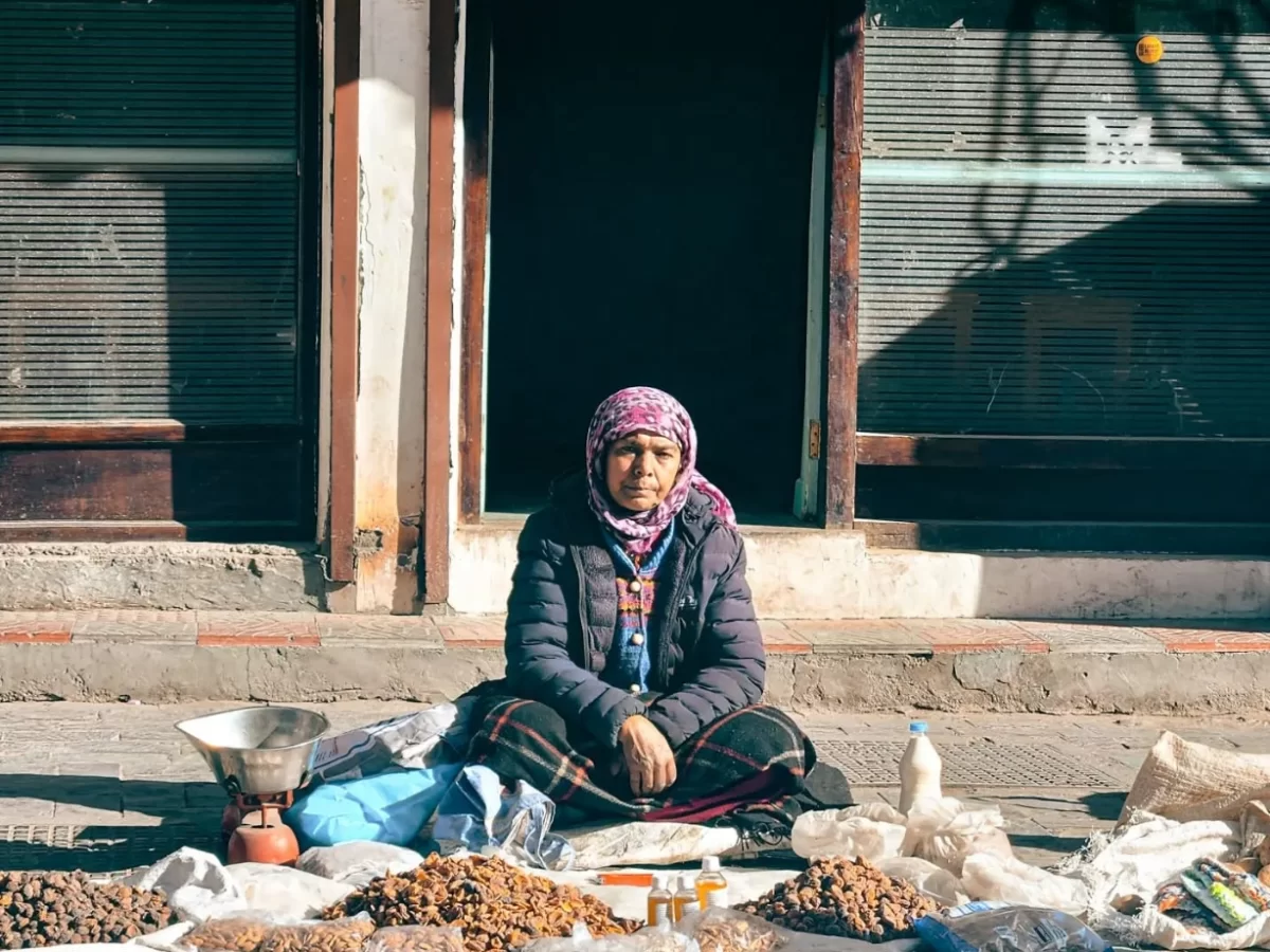 Elderly woman selling dry fruits and nuts seated outside shop in Leh Market, traditional attire amid Ladakhi architecture, perfect India tour package.