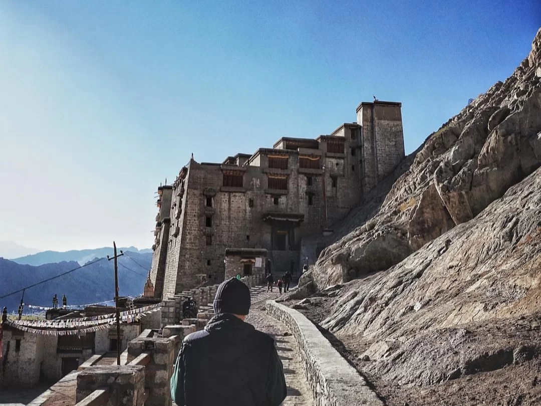 Leh Palace Ladakh dramatic multi-tiered white stone fortress towering over rocky terrain path with silhouetted hiker visitors distant snowy mountains blue sky, epic Himalayan palace trek tour package.