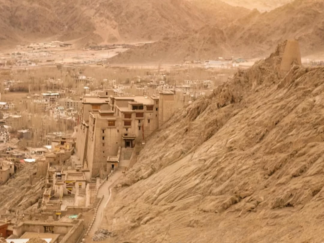 Leh Palace Ladakh golden hour panoramic view of ancient multi-storeyed beige stone palace ruins perched on arid hill overlooking Leh town rugged mountains hazy atmosphere, timeless Himalayan heritage tour package.