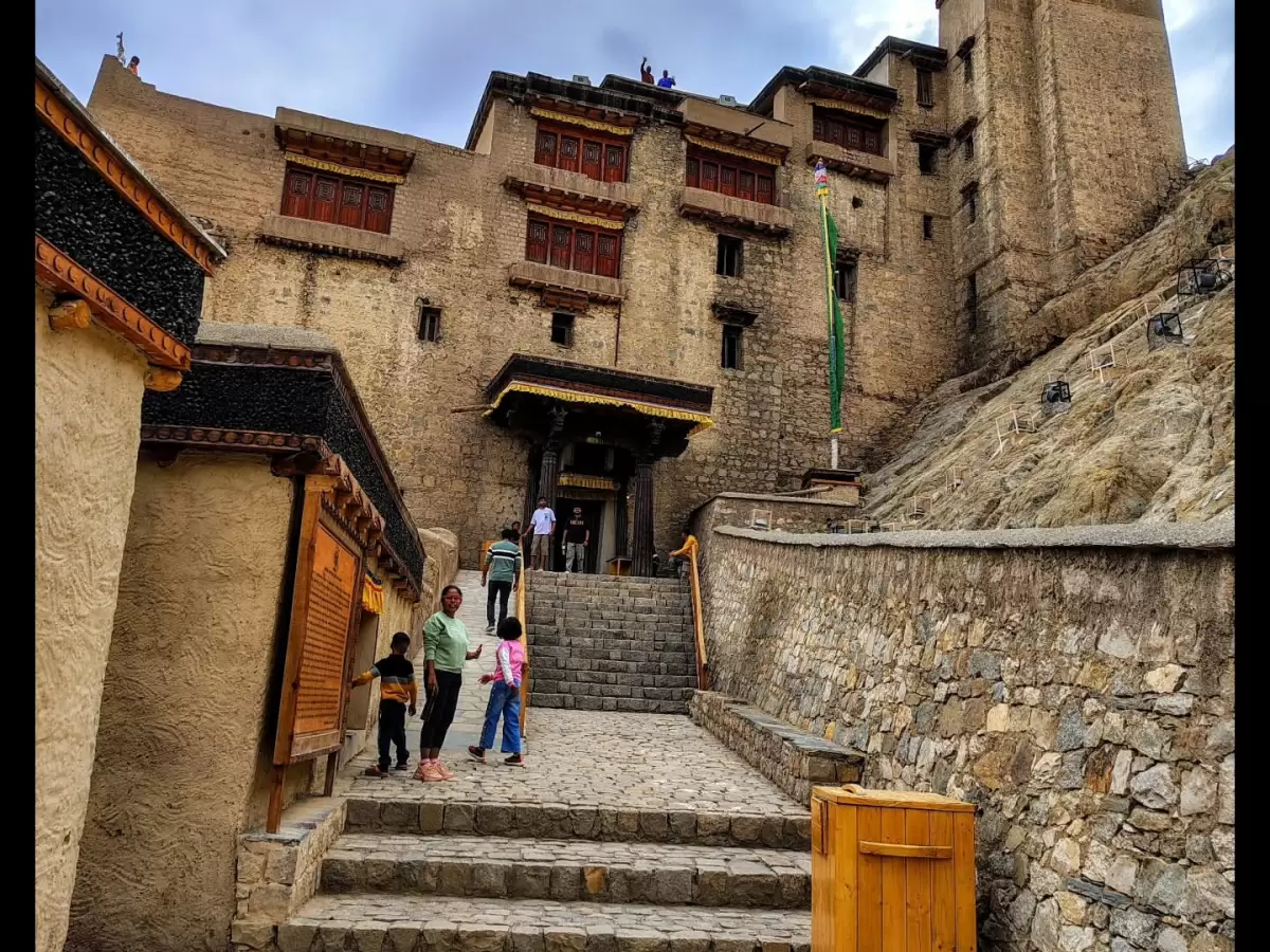 Leh Palace Ladakh close-up tourists family ascending stone steps to grand beige stone entrance with red wooden doors fluttering prayer flags cloudy sky backdrop, vibrant Himalayan royal heritage tour.