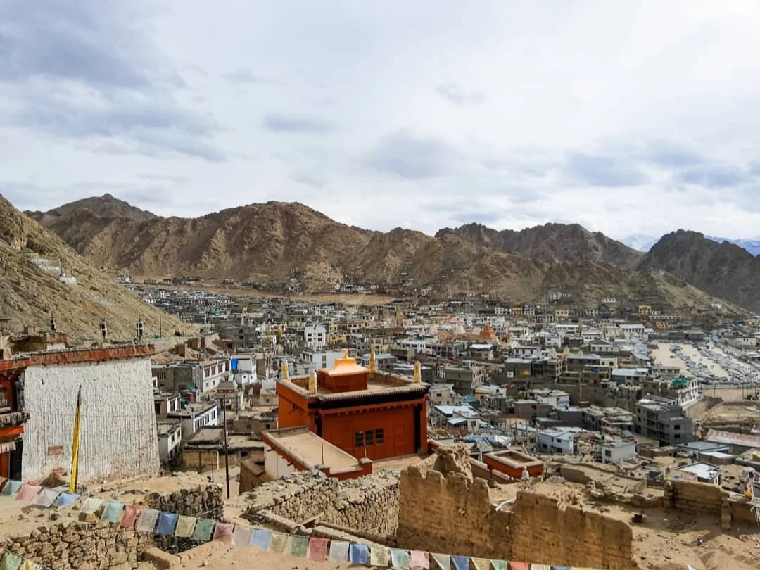 Leh Palace Ladakh elevated panoramic view over traditional Ladakhi town with colorful prayer flags red roofed buildings ruins against dramatic rugged mountains cloudy skies, immersive Himalayan cultural adventure tour package.