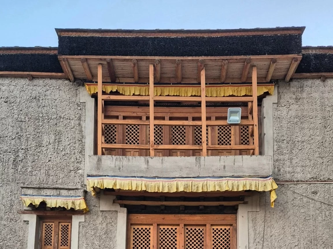 Leh Palace ornate wooden balcony with latticed panels draped in yellow prayer flags, flanked by carved doors on textured mud-brick wall under pale blue sky, perfect India tour package