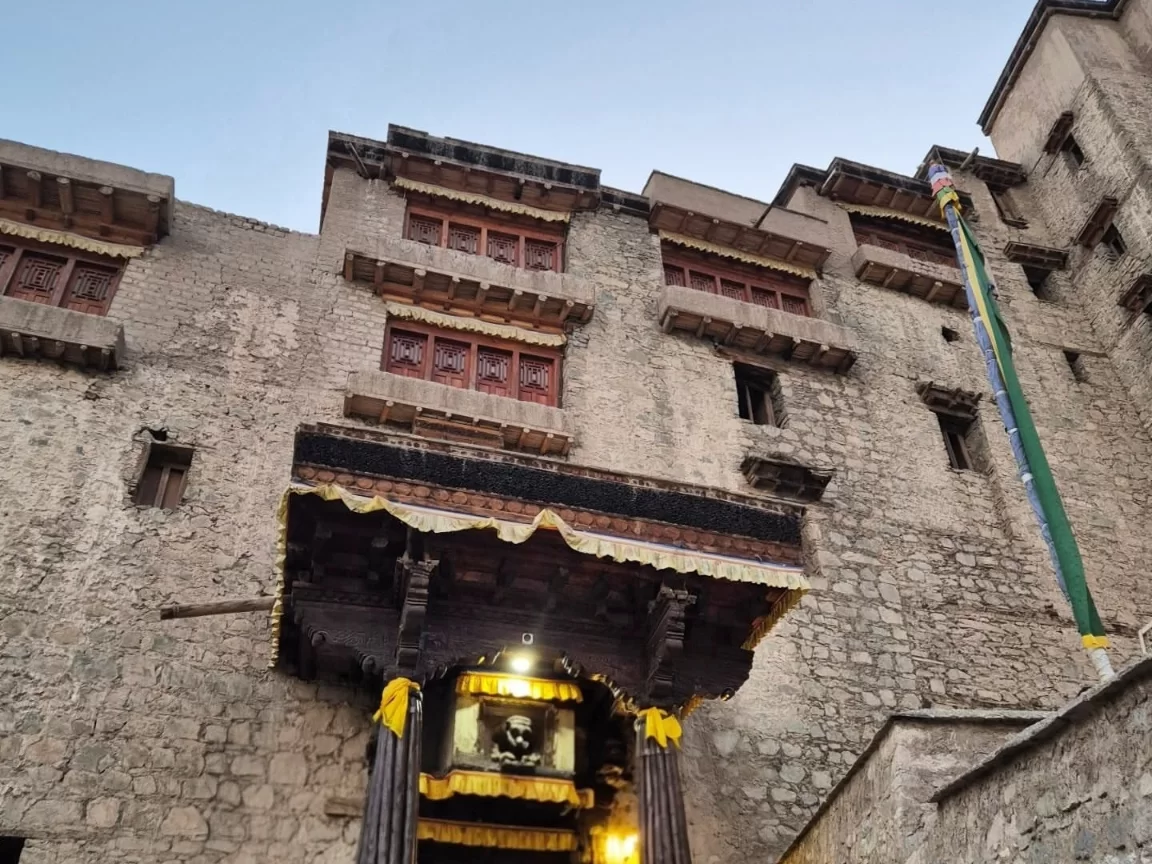 Leh Palace grand entrance with gilded framed portrait under ornate yellow-trimmed wooden canopy, flanked by red-framed windows on rugged stone walls against twilight sky, perfect India tour package.