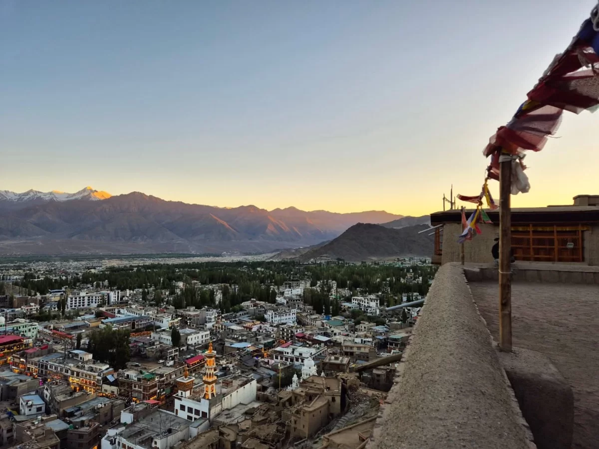Leh Palace terrace view over Leh town with colorful buildings along Indus Valley, snow-capped Stok Kangri mountains at sunset, fluttering prayer flags in foreground, perfect India tour package.