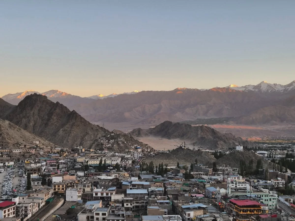 Leh Palace panoramic view of Leh city sprawled in Indus Valley with white and red-roofed houses amid poplars, rugged hills and distant snow peaks glowing at golden hour sunset, perfect India tour package.