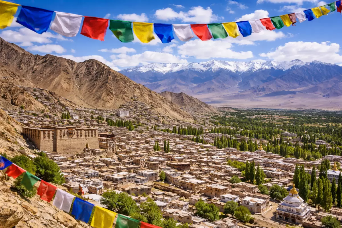 A panoramic view of Leh city and the historic Leh Palace under a bright blue sky, featured in a Leh Ladakh budget tour package, with colorful Tibetan prayer flags in the foreground and snow-capped Himalayan peaks in the distance.