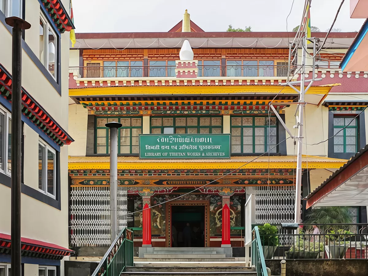 Library of Tibetan Works and Archives in Dharamshala featuring a traditional Tibetan-style building with colorful wooden carvings, green signboard at the entrance, and decorative pillars leading up a short staircase.