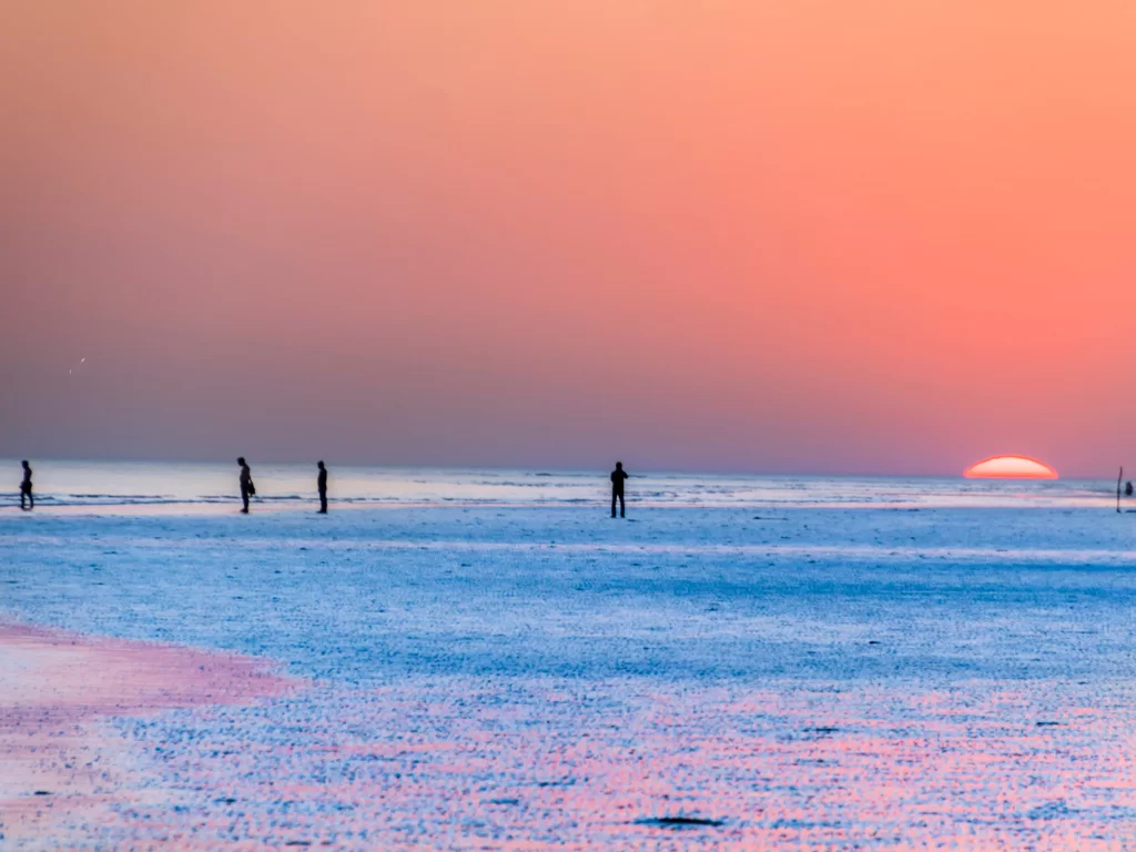 Mandvi Beach sunset at Kutch Gujarat during golden hour, featuring silhouetted people, orange-pink skies, glowing sun on white sands and sea waves, perfect romantic experience with Gujarat tour packages.