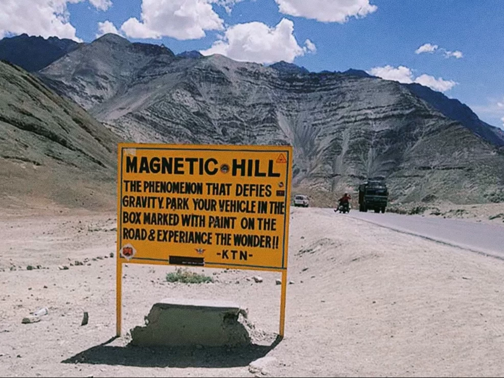 Magnetic Hill sign at Ladakh during partly cloudy day, featuring gravity phenomenon board rugged mountains road, perfect adventure Ladakh tour package.
