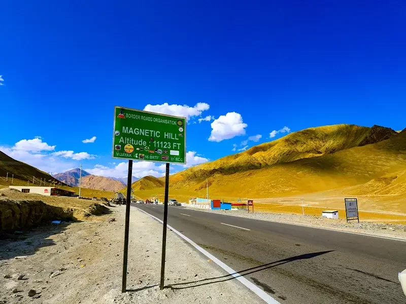 Magnetic Hill sign at Ladakh BRO during clear skies, featuring altitude 11312 ft board yellow mountains highway, perfect adventure Ladakh tour package. 