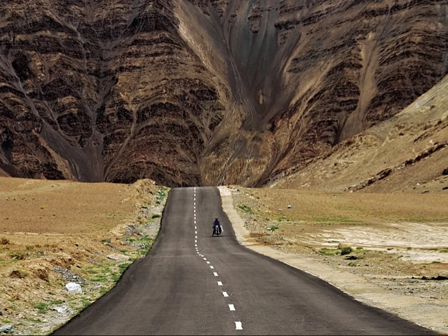 Magnetic Hill Ladakh Leh-Kargil highway dramatic barren valley towering rugged Zanskar mountains lone motorcyclist straight asphalt road sparse desert shrubs hazy horizon, famous optical illusion gravity hill tourist attraction adventure tour package.