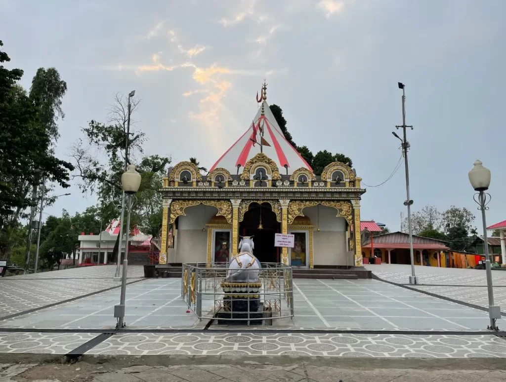 Mahabhairab Temple Tezpur during golden hour, featuring colorful striped temple dome gold ornate entrance white Shiva statue courtyard lamps trees, perfect cultural heritage Tezpur Assam tour package.