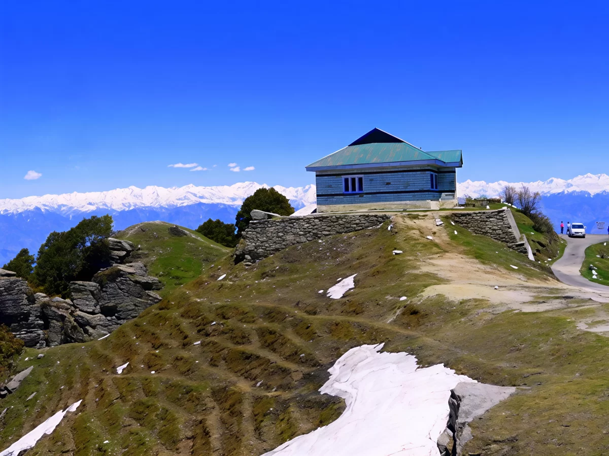 Mahasu Peak in Kufri featuring a small hilltop building with green roof, winding mountain road, grassy slopes with patches of snow, and panoramic snow-capped Himalayan ranges under a clear blue sky.