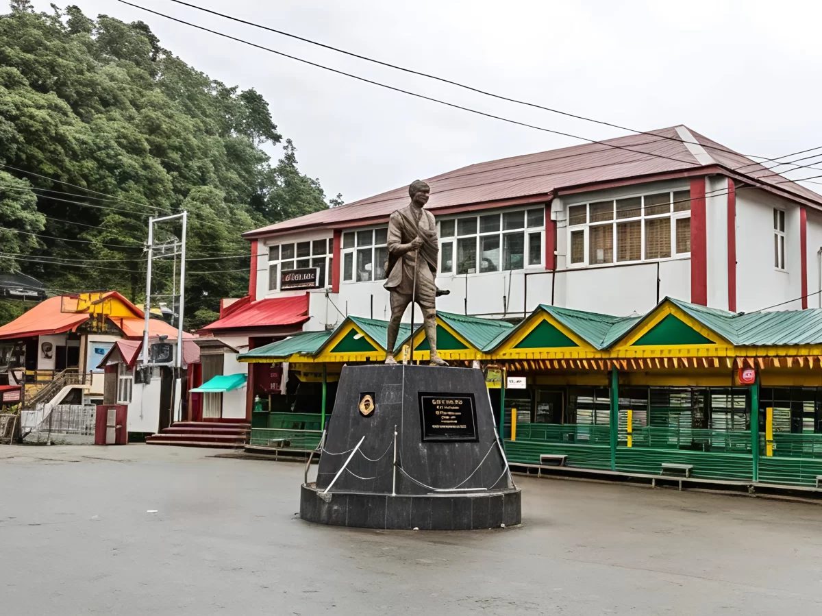 Mall Road in Dalhousie featuring a central statue of Mahatma Gandhi on a pedestal, surrounded by colorful shopfronts and colonial-style buildings under an overcast sky.