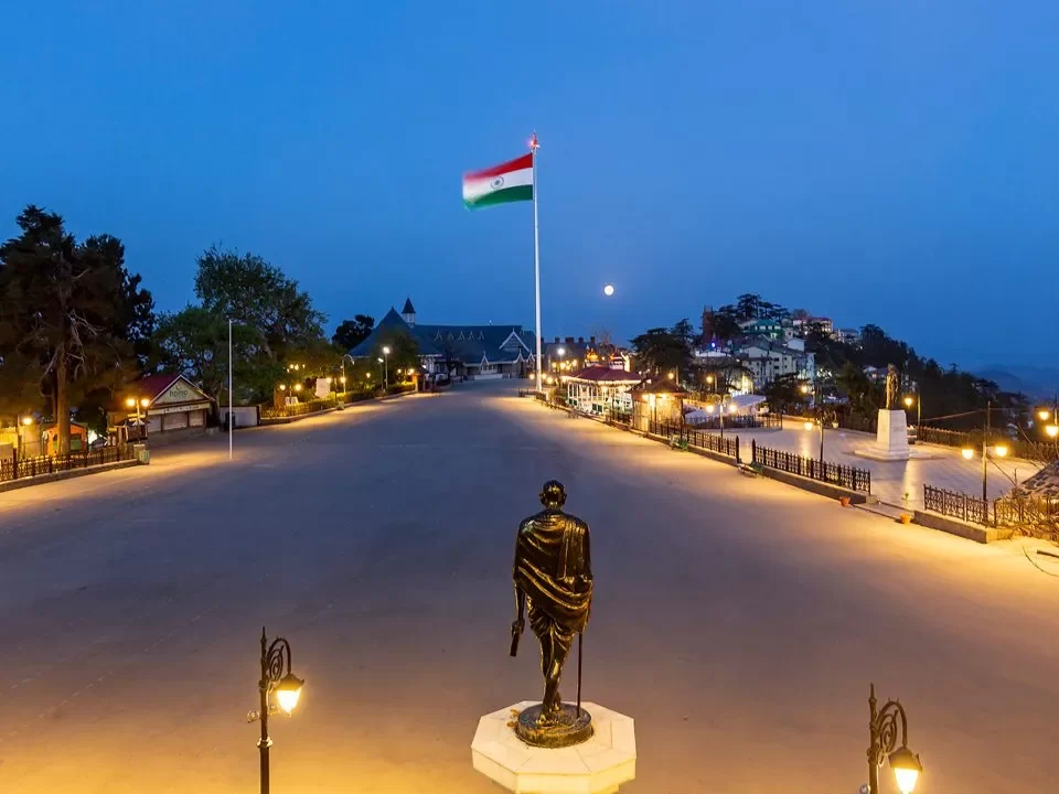 Gandhi Statue at Mall Road Shimla during twilight, featuring Indian flag, colonial buildings, lamps, hills, perfect evening stroll, Himachal Pradesh tour packages.
