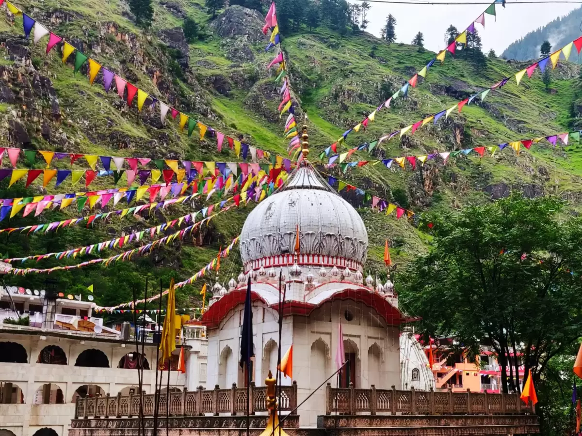 Manikaran Sahib Gurudwara Parvati Valley near Kasol Himachal Pradesh, featuring white dome temple complex colorful prayer flags green forested rocky mountain backdrop saffron flags foreground, perfect cultural experience Himachal tour package.