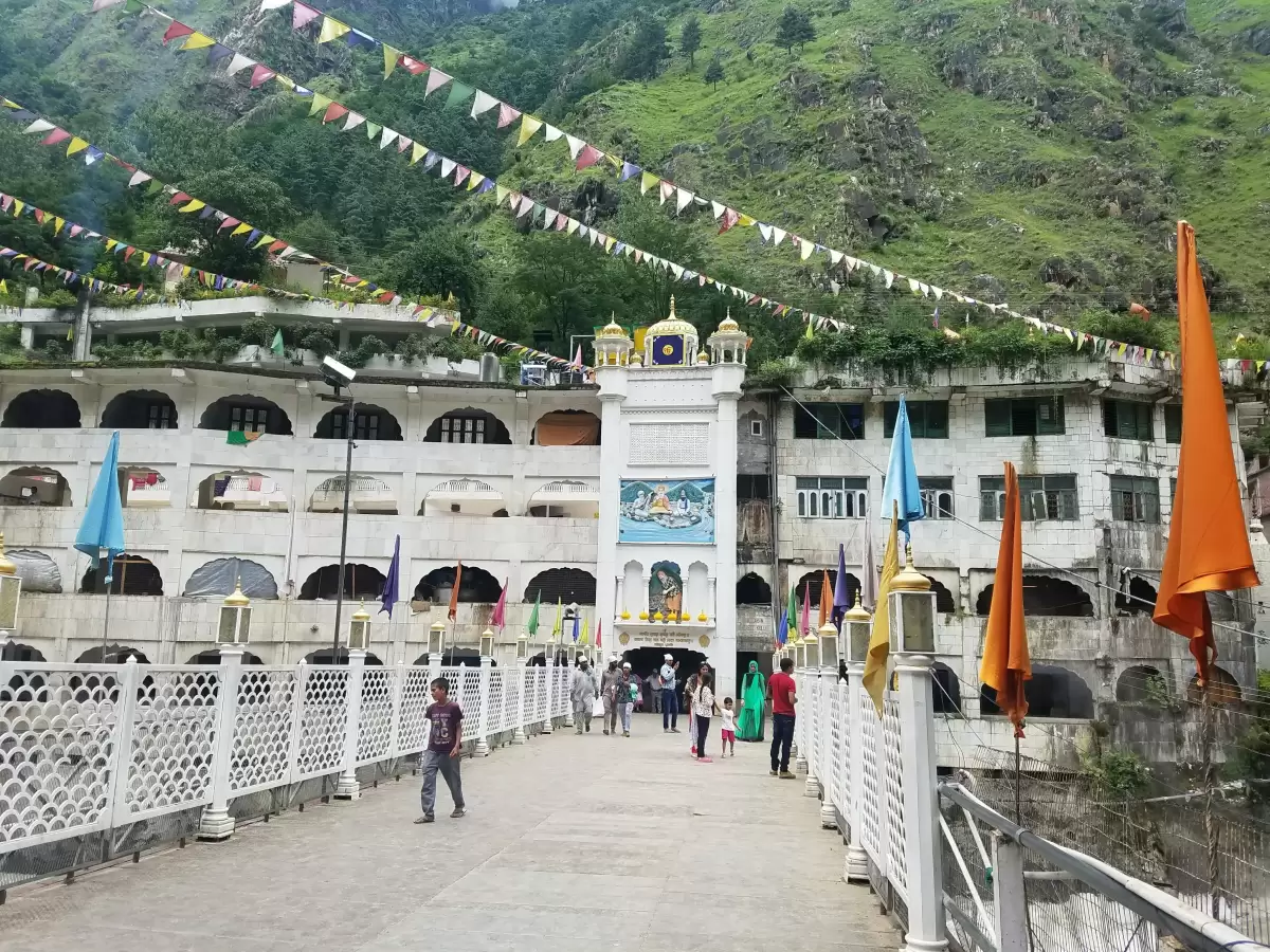 Manikaran Sahib Gurudwara Parvati Valley near Kasol Himachal Pradesh, featuring white arched temple complex clock tower prayer flags green forested mountain backdrop visitors bridge foreground, perfect cultural experience Himachal tour package.