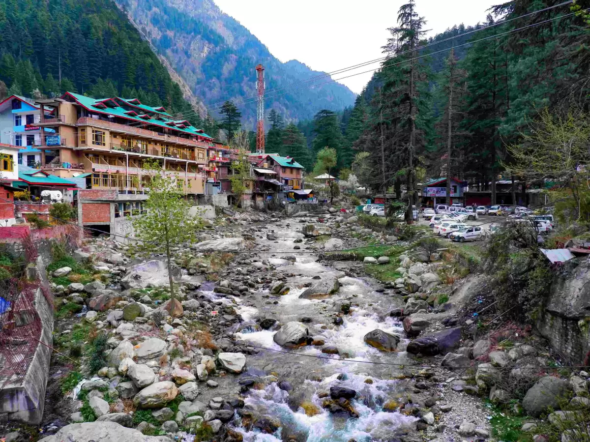 Manikaran town Parvati Valley near Kasol Himachal Pradesh during overcast skies, featuring colorful multi-story hotels guest houses parked vehicles rocky Parvati River pine forested mountain backdrop, perfect cultural experience Himachal tour package.