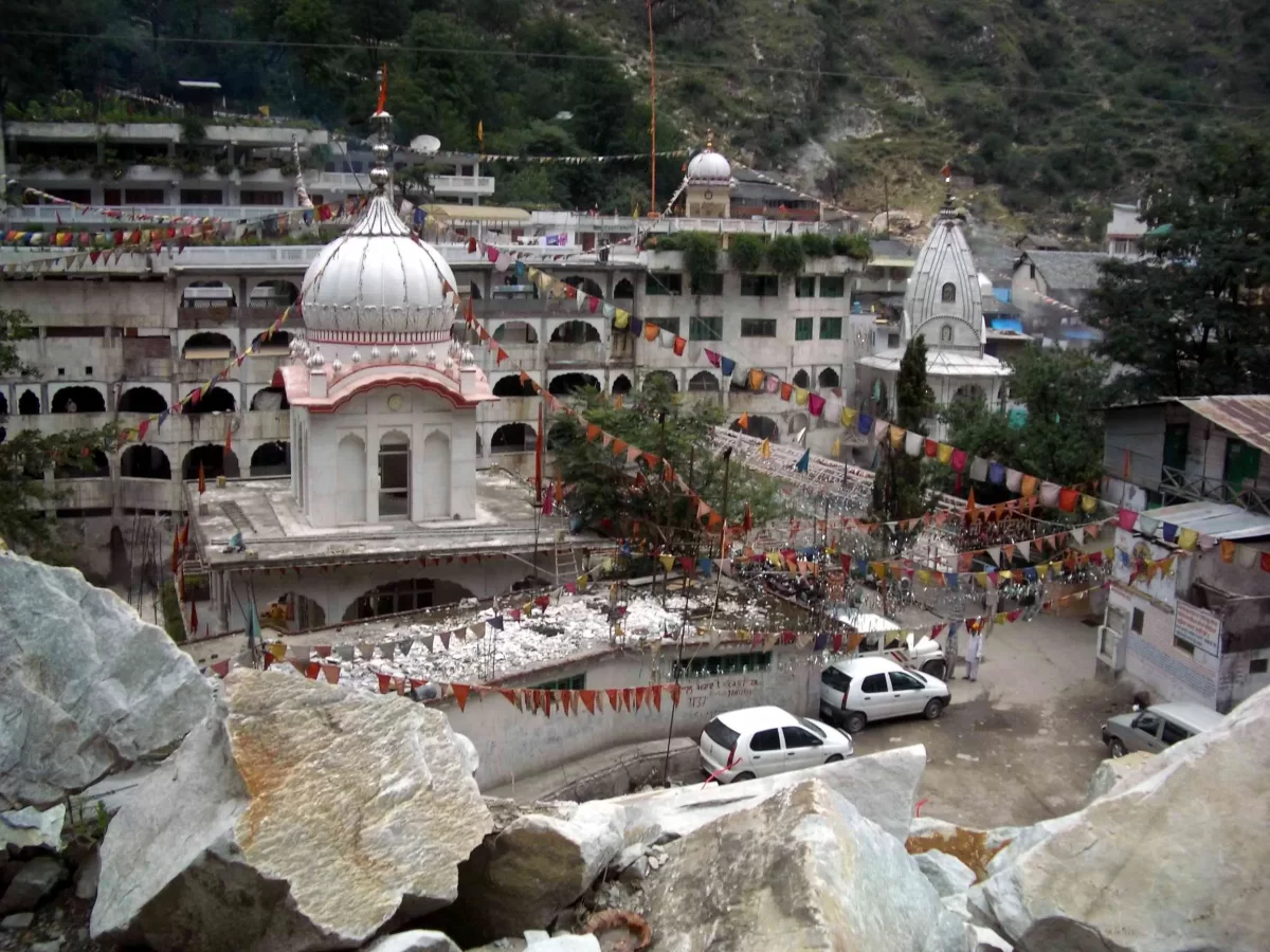 Manikaran Sahib Gurudwara Parvati Valley near Kasol Himachal Pradesh, featuring white domed temples prayer flags complex rocky Parvati River backdrop vehicles foreground, perfect cultural experience Himachal tour package.