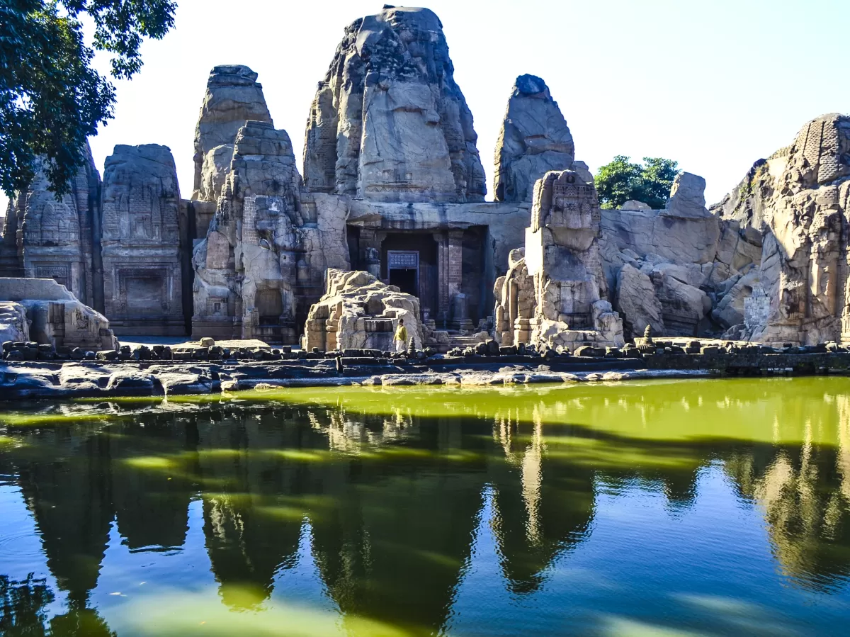 Masroor Rock-Cut Temple near Dharamshala featuring ancient monolithic stone temples carved from rock, reflected in a green water tank with clear blue sky above.