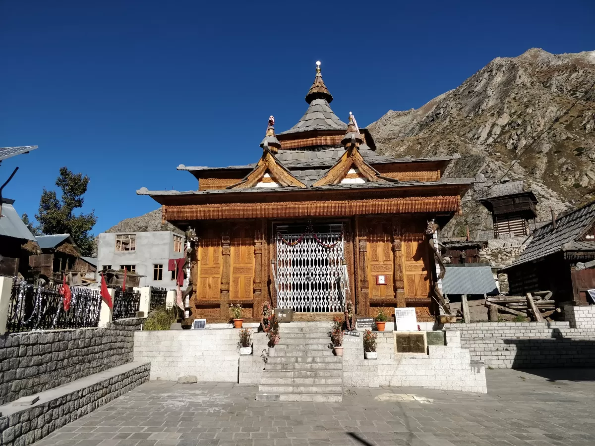 Mathi Temple Chitkul Kinnaur Himachal Pradesh during clear blue skies, featuring ornate wooden pagoda temple carved doors snowy mountains background village huts, perfect cultural experience Himachal tour package.