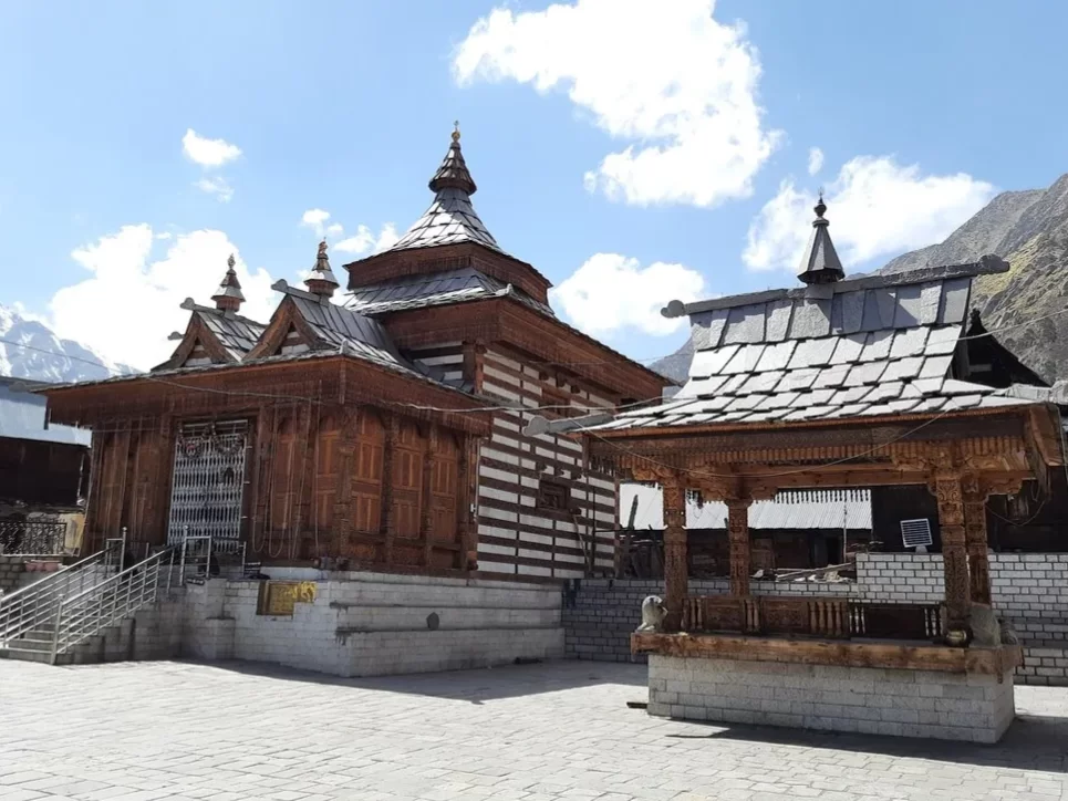 Mathi Temple in Chitkul, Kinnaur Himachal Pradesh during partly cloudy blue skies, featuring ornate wooden pagoda-style temple intricate carvings snowy peaks background, perfect cultural experience Himachal tour package.
