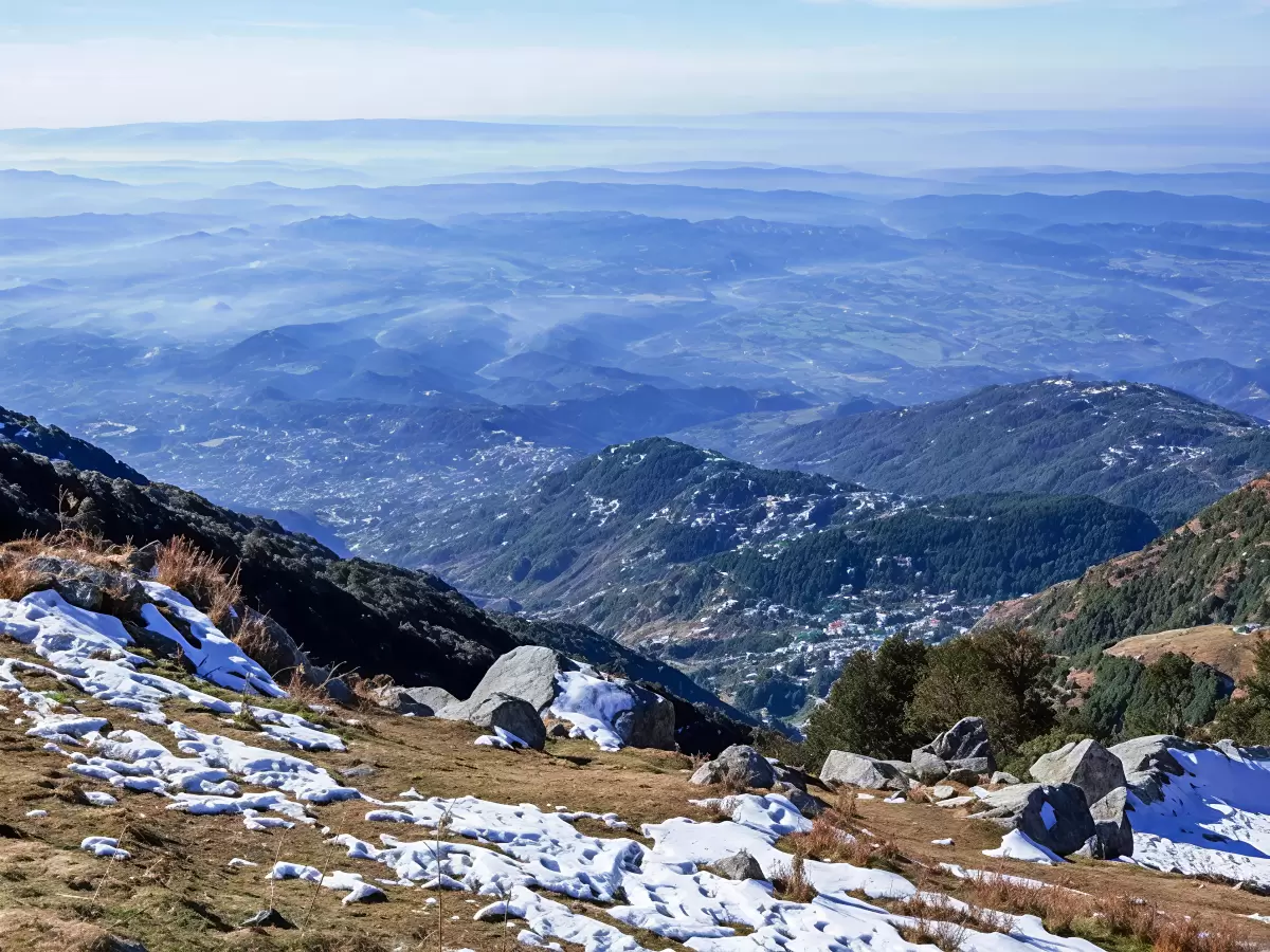 McLeod Ganj Dharamshala Himachal Pradesh during clear hazy skies, featuring panoramic Dhauladhar mountain ranges valley townships snow patches rocky meadows foreground, perfect cultural experience Himachal tour package.