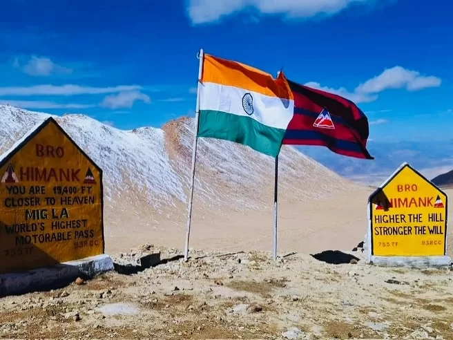 BRO signboards with flags at Mig La Ladakh during clear sunny day, featuring Himank world's highest motorable pass plaques, Indian tricolor, snowy peaks desert terrain, perfect adventure Ladakh tour package.