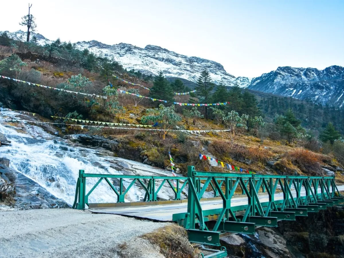 Green bridge over Khanda waterfall at Katao Lachung Sikkim amid prayer flags and snow peaks, perfect adventure North Sikkim tour package.