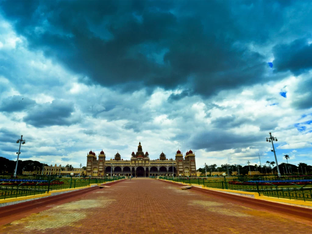 Mysore Palace under dramatic monsoon clouds, featuring grand facade, central walkway and lush gardens, perfect Karnataka tour package