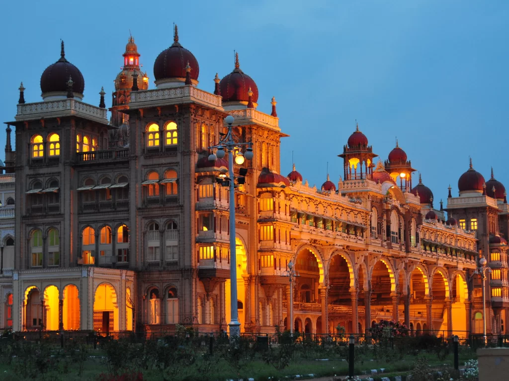 Mysore Palace illuminated at dusk, featuring glowing arches, domes and landscaped gardens, perfect royal Karnataka tour package