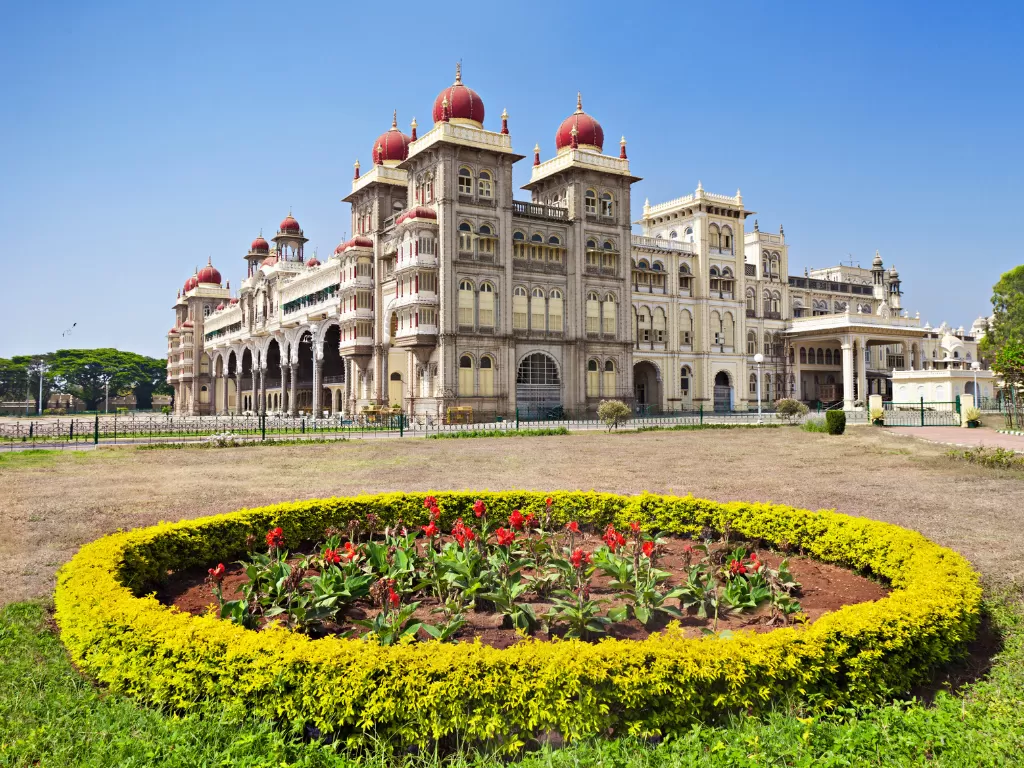 Mysore Palace in daytime, featuring circular flower garden, arched facade and red domes under blue sky, perfect royal Karnataka tour package