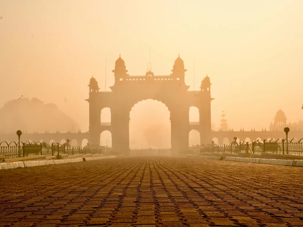 Foggy morning view of Mysore Palace gateway, featuring grand archway, cobbled path and soft golden light, perfect Karnataka tour package