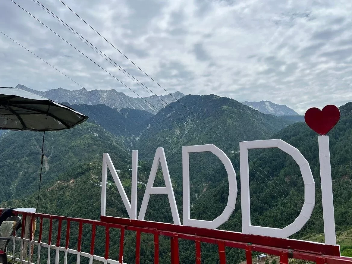 Naddi View Point McLeod Ganj Dharamshala Himachal Pradesh during cloudy skies, featuring Naddi signboard Dhauladhar snow-capped mountain range pine forested hills backdrop umbrella railing foreground, perfect cultural experience Himachal tour package.