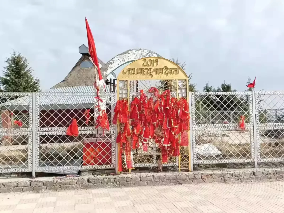 Entrance of Nag Temple in Kufri featuring a decorative golden archway adorned with red prayer flags, white metal fencing, and temple structure in the background under a cloudy sky.