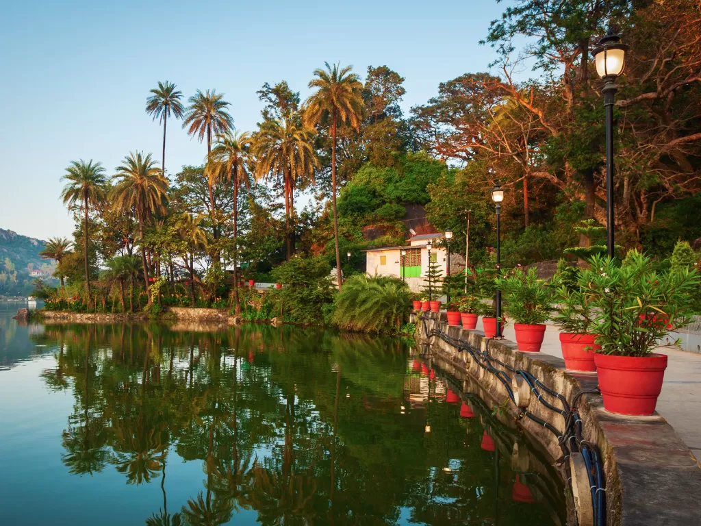 Nakki Lake Mount Abu lakeside promenade, featuring palm trees potted plants lamps white buildings reflecting turquoise waters green hills blue skies, perfect Rajasthan tour packages.
