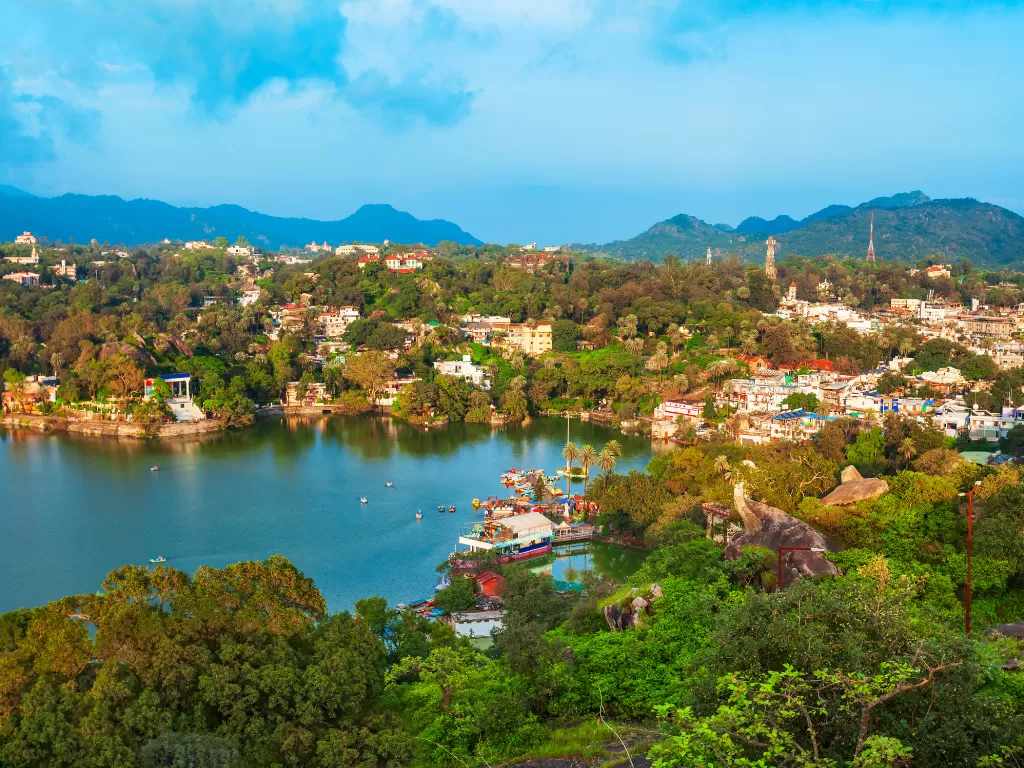 Nakki Lake Mount Abu aerial vista, featuring turquoise waters boats houseboats surrounding green hills town buildings distant mountains cloudy blue skies, perfect Rajasthan tour packages.