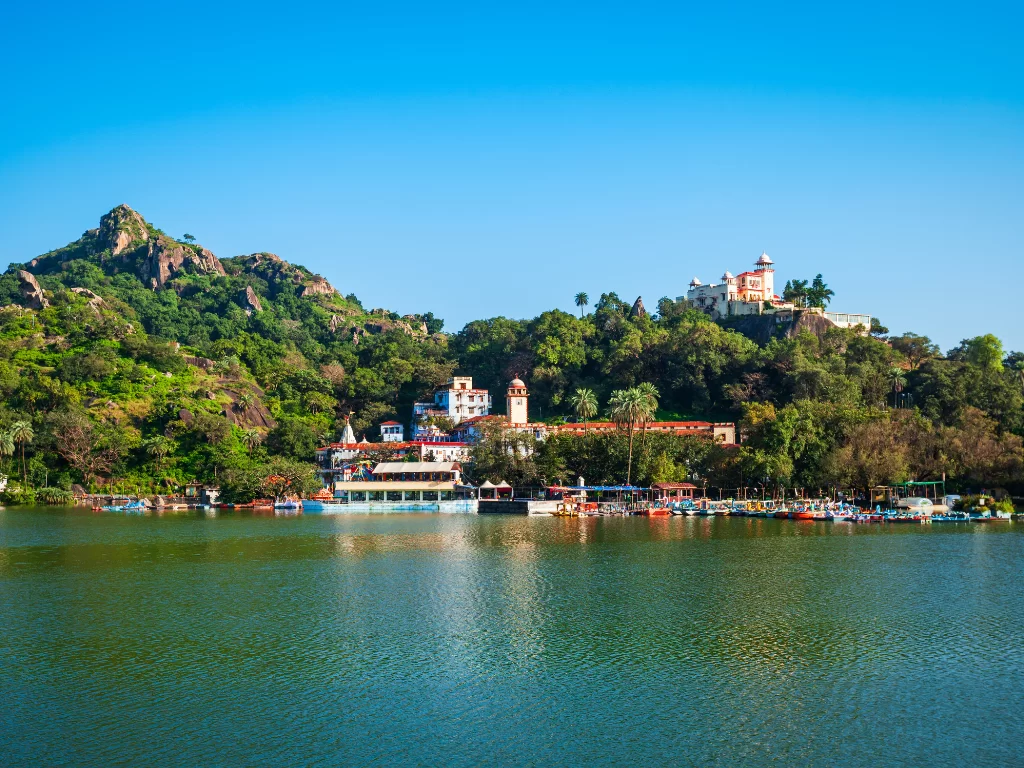 Nakki Lake Mount Abu scenic view, featuring turquoise lake colorful boats houseboats palace hotels palm trees rocky hills lush greenery blue skies, perfect Rajasthan tour packages.