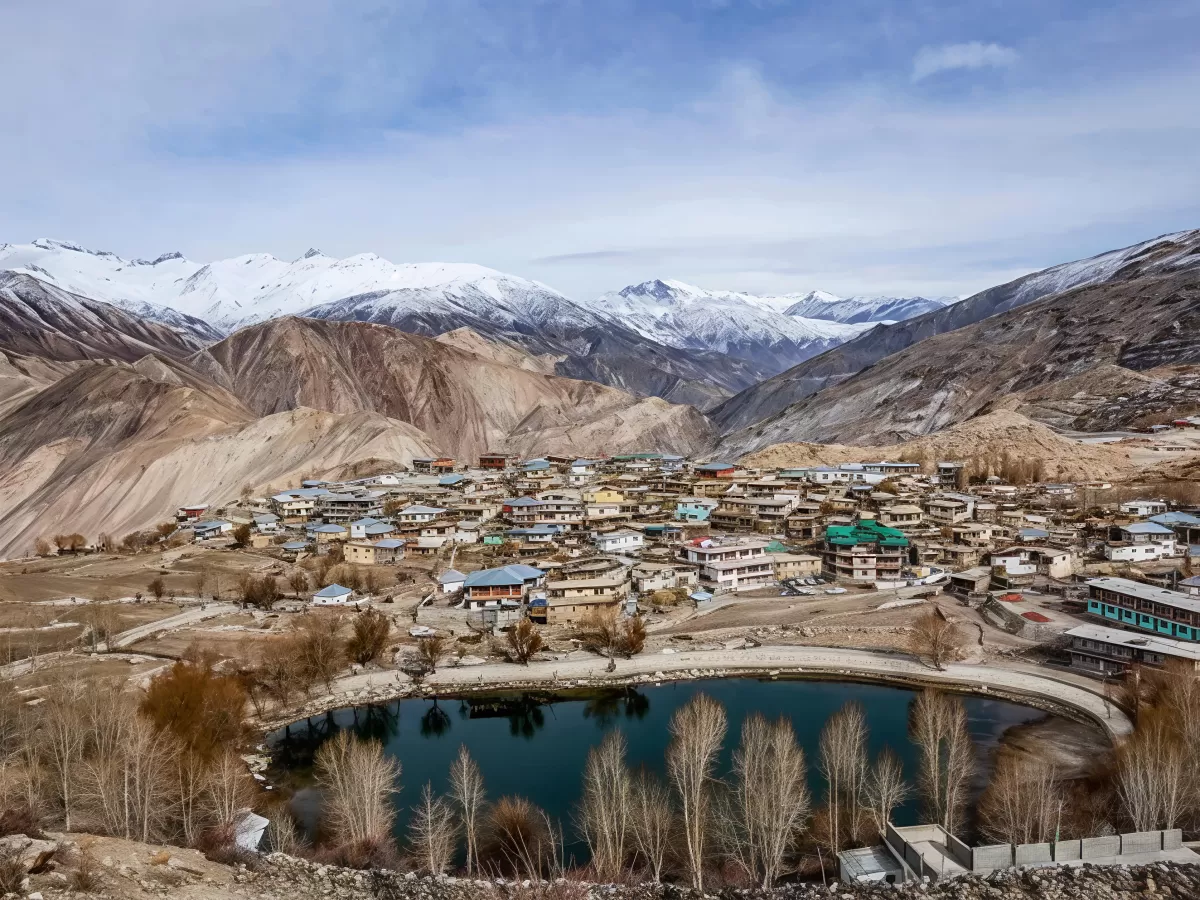 Nako Lake Nako village Spiti Valley during winter partly cloudy skies, featuring dark blue waters bare willow trees colorful houses barren hills snow-capped mountains, perfect nature experience Himachal Pradesh tour package.