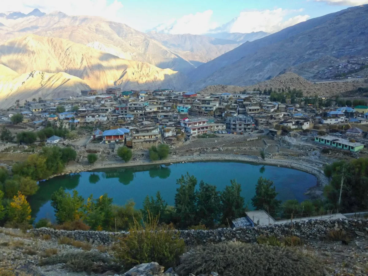 Nako Lake Nako village Spiti Valley during golden hour partly cloudy skies, featuring turquoise waters colorful clustered houses willow trees barren mountains snow peaks, perfect nature experience Himachal Pradesh tour package.