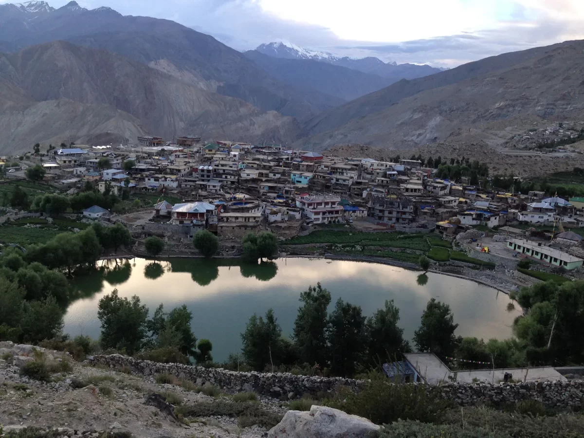 Nako Lake Nako village Spiti Valley during evening partly cloudy skies, featuring turquoise waters colorful houses willow trees snow mountains surrounding hills, perfect nature experience Himachal Pradesh tour package.